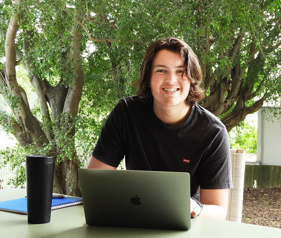 A young man sitting at an outdoor table with a laptop and notebook