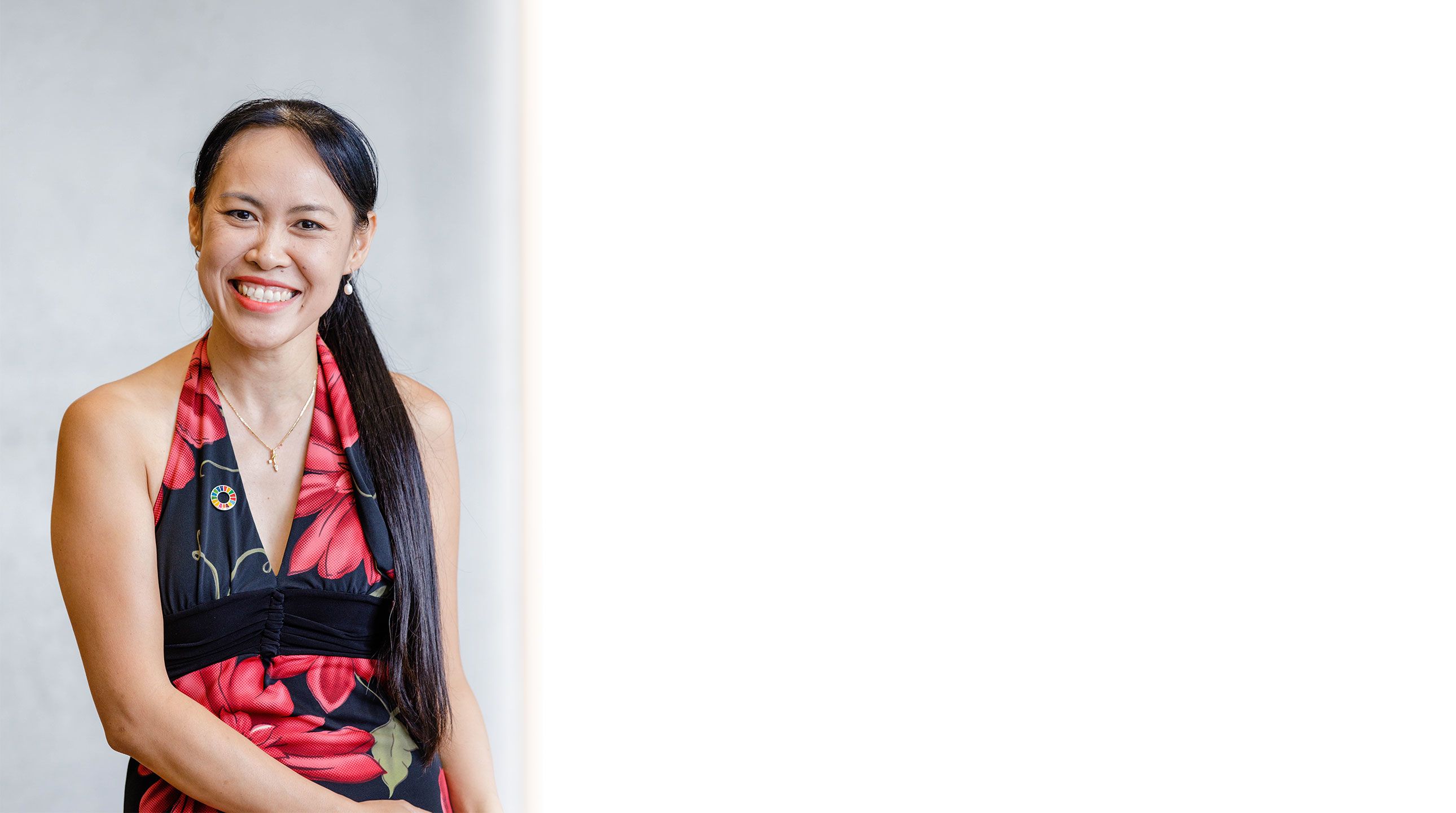 A woman with long dark hair and dark eyes smiles towards the camera. She is wearing white drop earrings and a red and white floral dress.