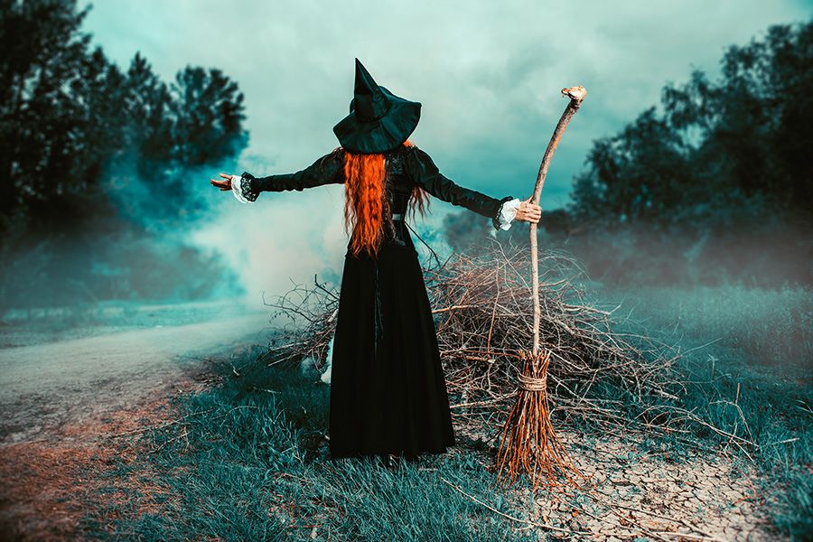 A person wearing a witch costume standing in a foggy clearing in a forest behind a pile of twigs. Their arms are outstretched and they are holding a broomstick.