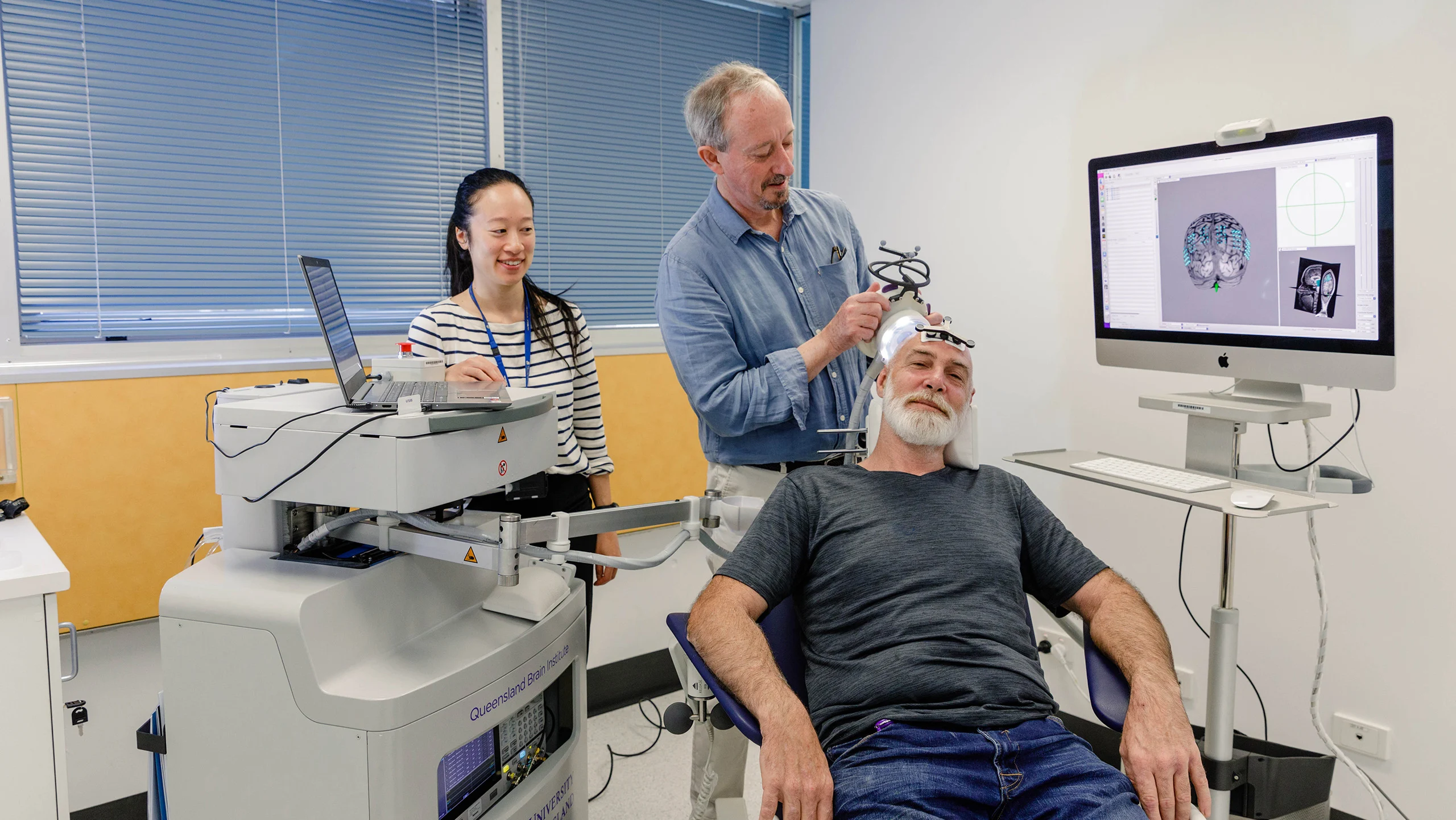 A man sitting on a chair receiving ultrasound treatment on his head in a lab. Two ultrasound technicians are behind him.