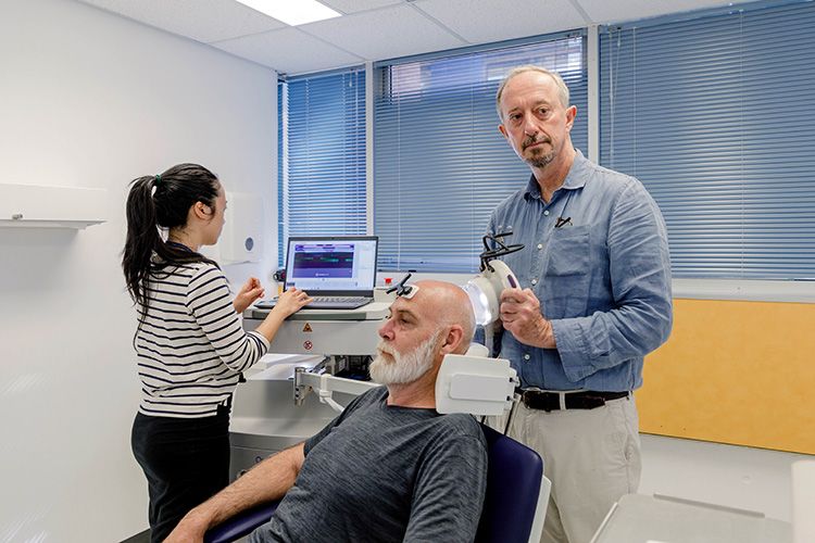 A man receiving ultrasound treatment on his head. Two technicians, one administering the ultrasound and one reading the results on a laptop, are behind the man.