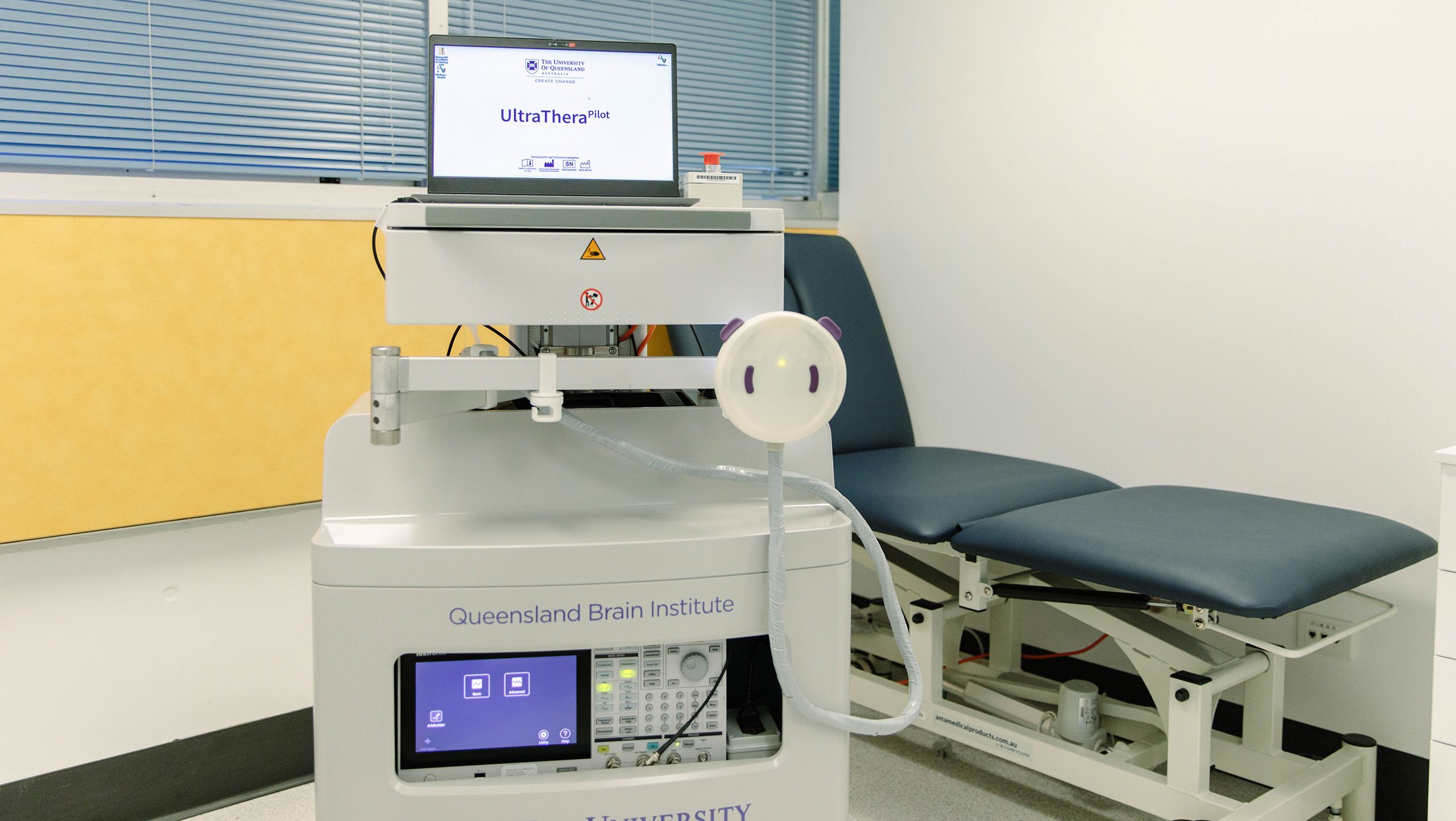 A laptop with a program called UltraThera Pilot open on its screen. The laptop is on top of a stand with an ultrasound device attached to it. A medical examination bed is in the background.