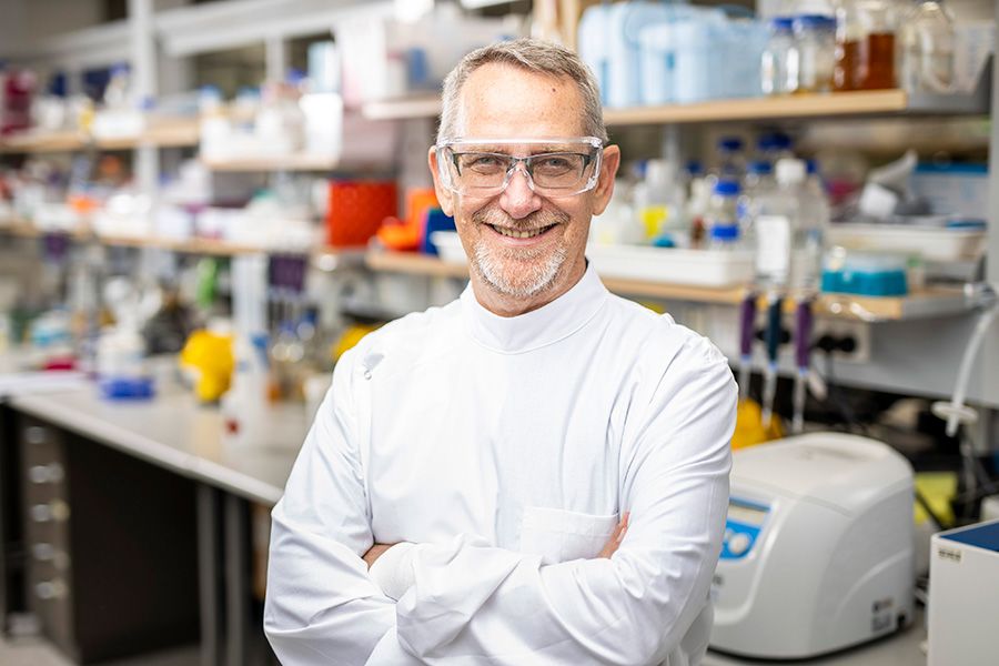 Professor Paul young wearing a lab coat and safety goggles. He is standing in a laboratory with his arms crossed and smiling.