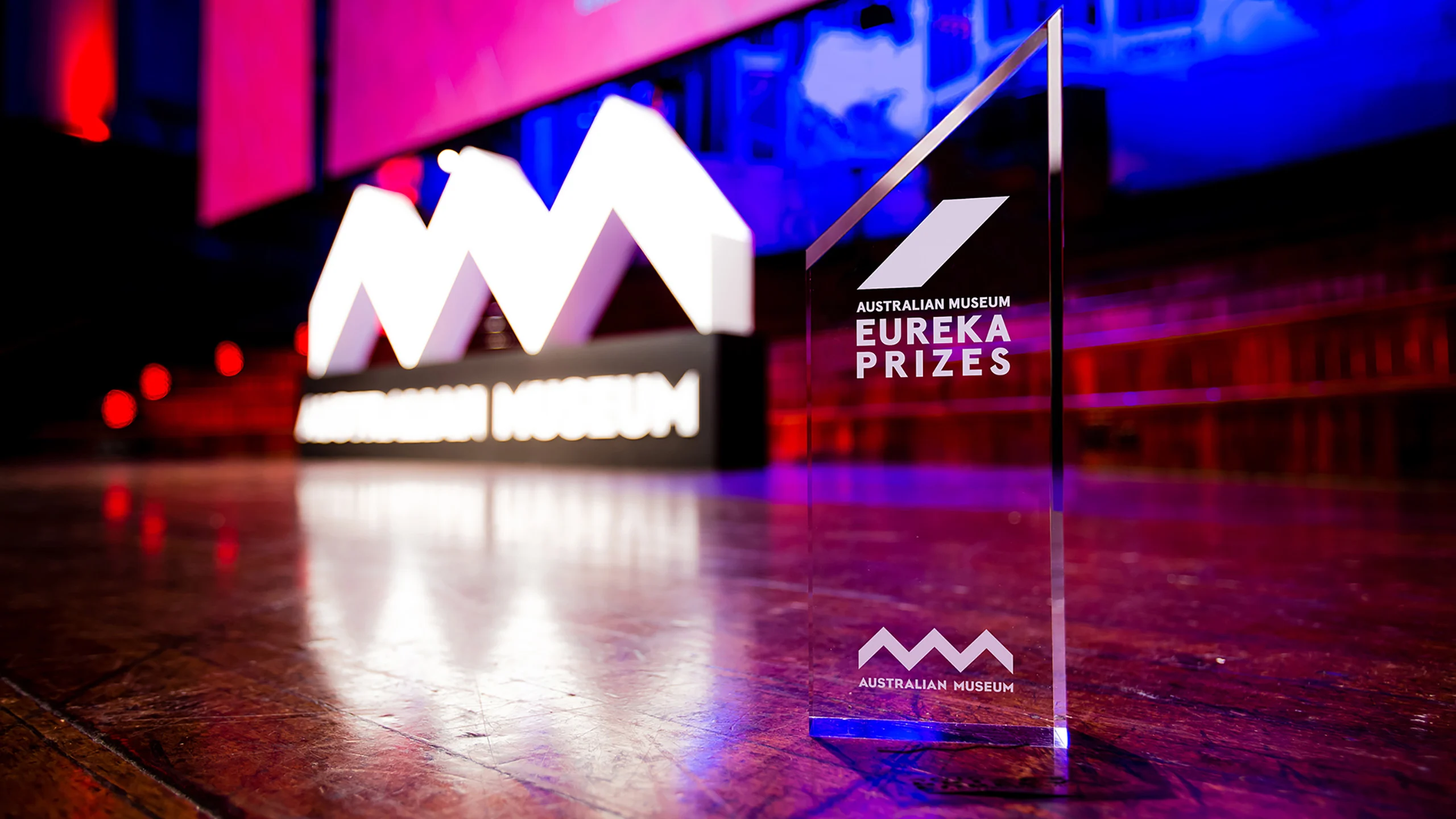 A glass trophy sitting on a wooden floor. Red and purple lights illuminate the room. There is a white sign reading Australian Museum in the background.