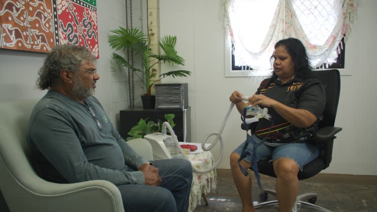 Two people sit in chairs facing each other, one is holding a plastic mask attached to a tube