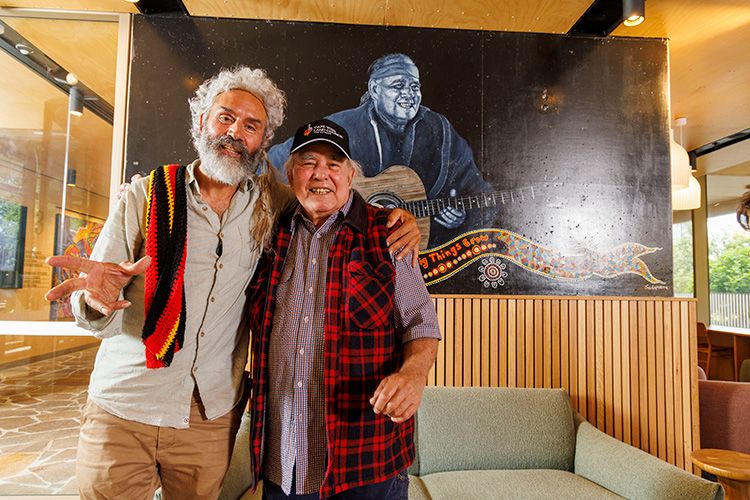 Dr Jandamarra Cadd, a middle-aged Indigenous man with grey hair, and Kev Carmody, an older Indigenous man, with their arms around each other's shoulders. They are standing in front of a mural of Kev playing the guitar.