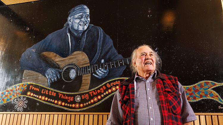 Kev Carmody, an older Indigenous man, smiling joyfully while standing in front of a mural of himself playing guitar