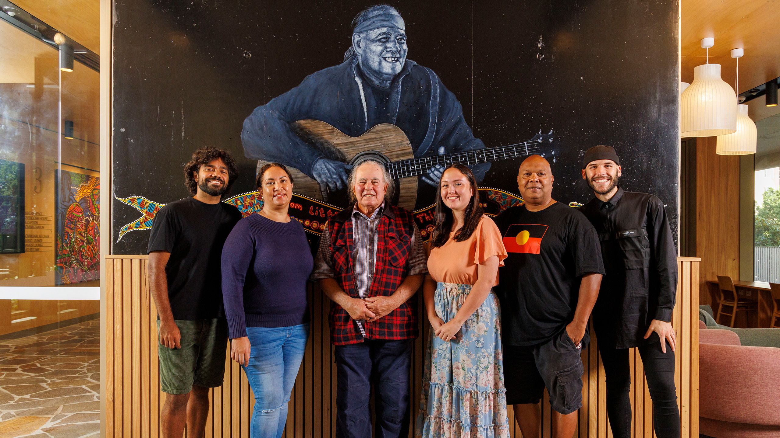 Kev Carmody, an older Indigenous man, with five younger Indigenous people standing in front of a mural of Kev playing guitar