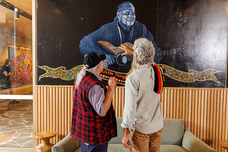 Kev Carmody and Dr Jandamarra Cadd examining a mural of Kev playing guitar. They both have their backs turned.