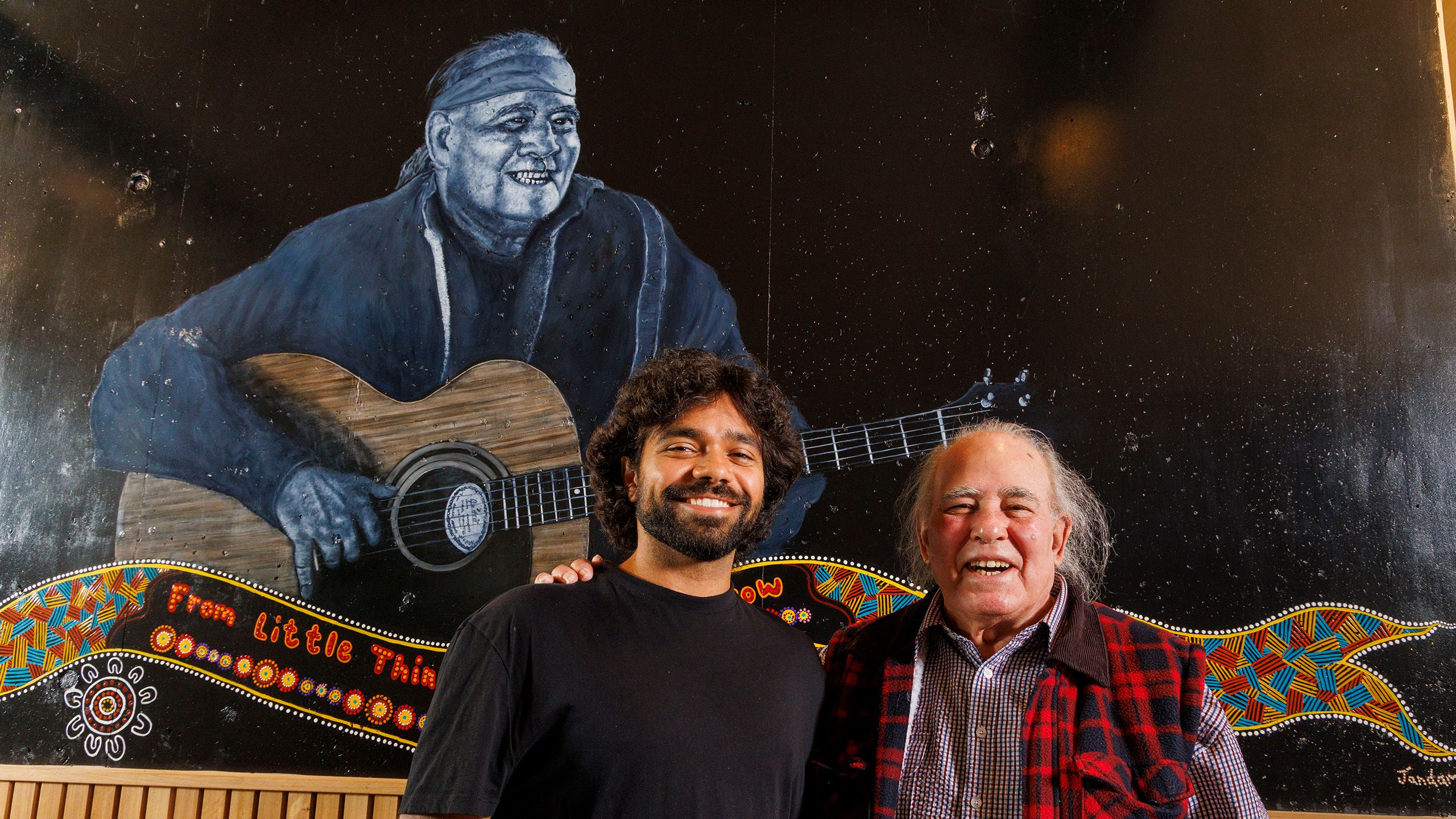 Thomas Scanlan, a young Indigenous man, with Kev Carmody, an older Indigenous man. They are standing in front of a large mural of Kev playing guitar.