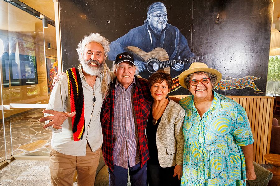 Dr Jandamarra Cadd, Kev Carmody, Professor Bronwyn Fredericks and Professor Tracey Bunda with their arms around each other and smiling. They are standing in front of a mural of Kev playing guitar.