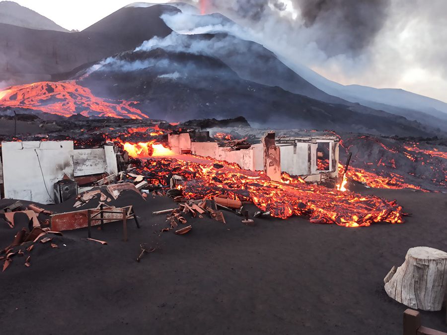 Pieces of buildings and furniture covered in volcanic ash and magma. A smoking volcano is behind the buildings and magma