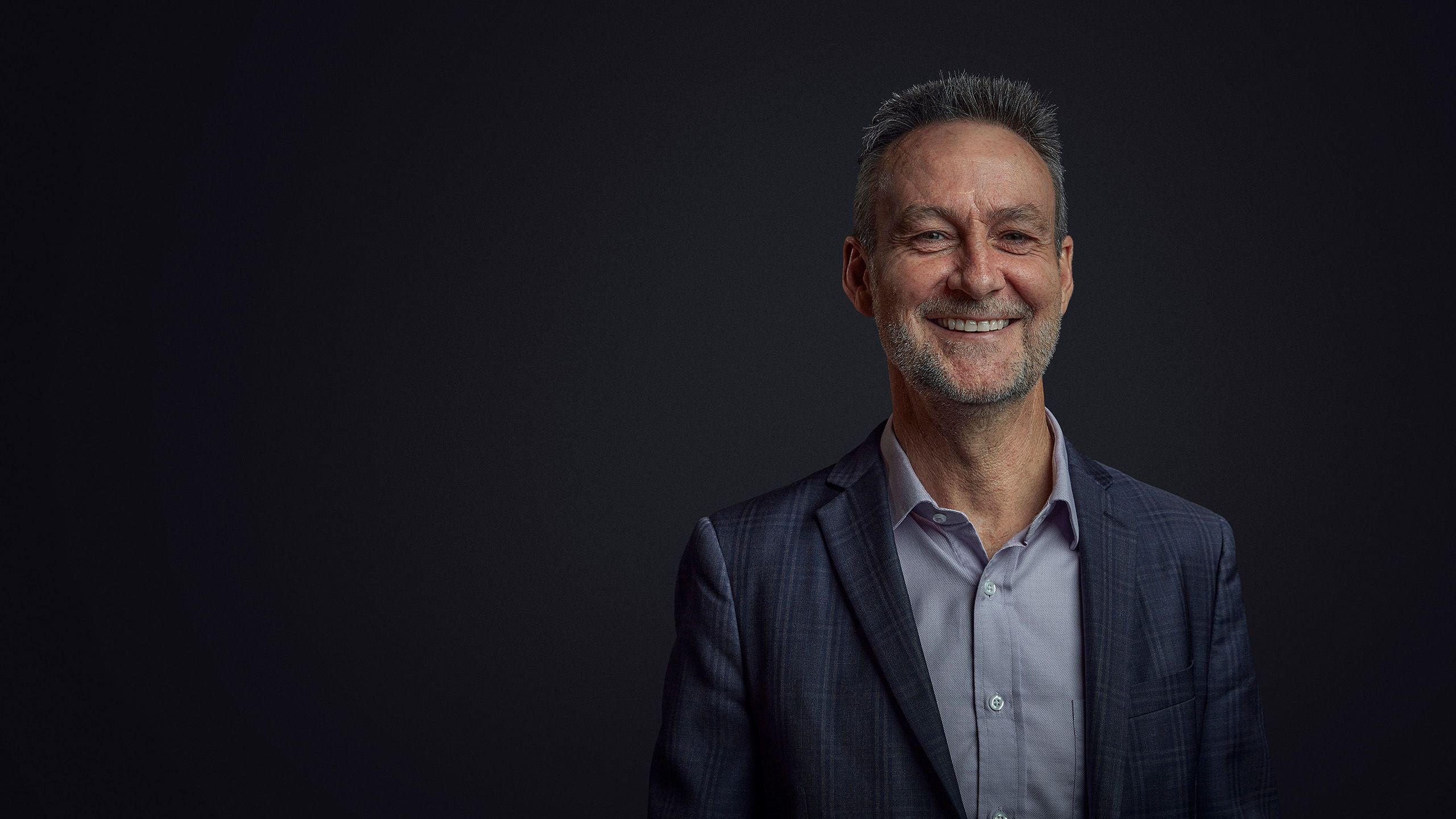 Glenn King wearing a jacket and collared shirt smiling broadly in front of a black background