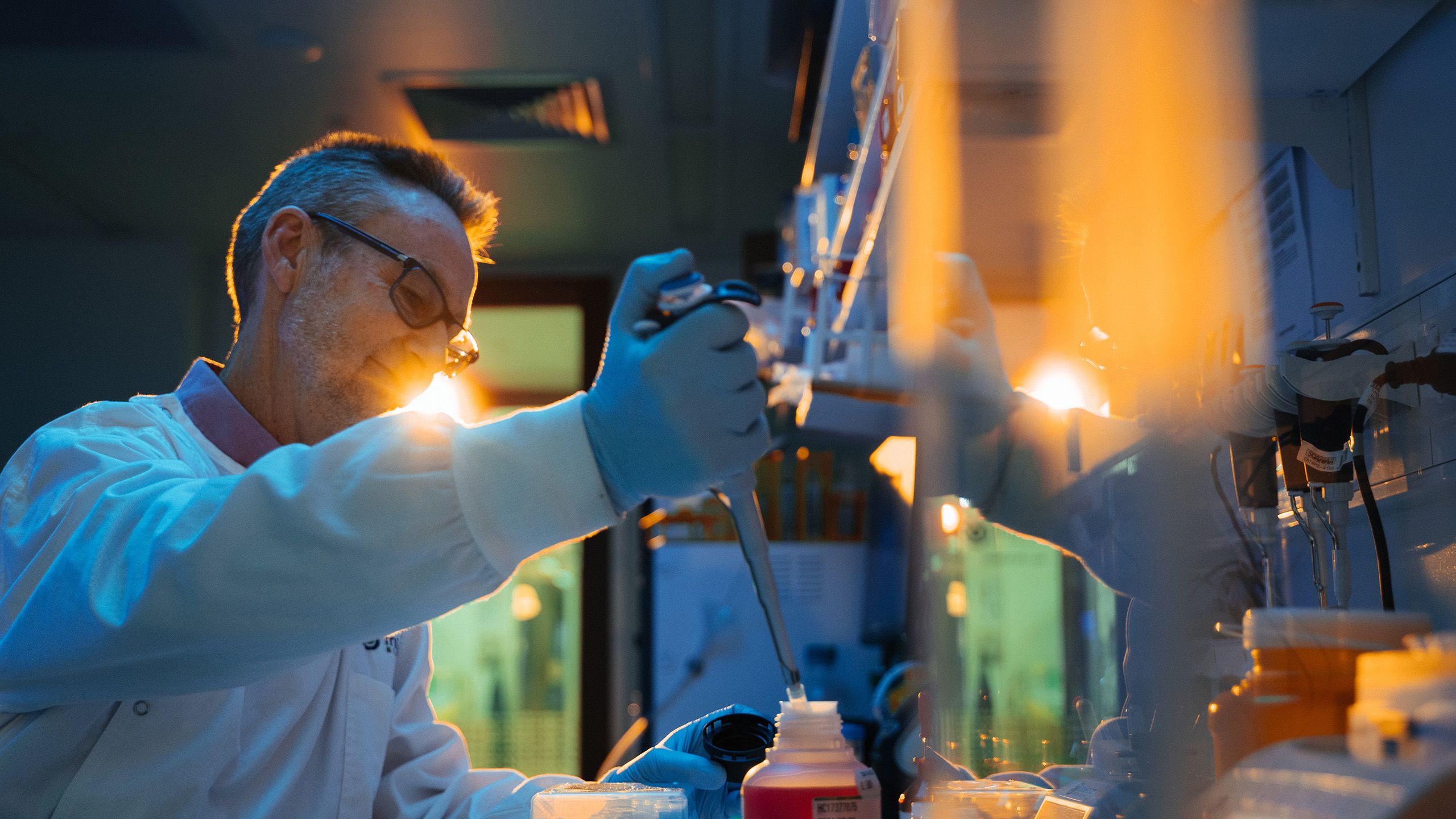 Professor Glenn King transferring liquid into a bottle with a pipette while standing in a lab. A bright light near the lab door is shining on his face.