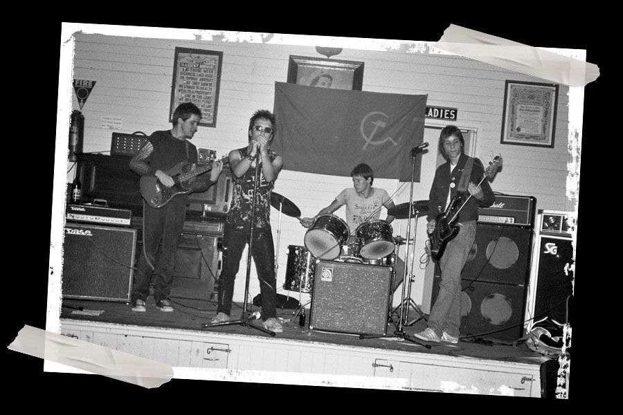 A black and white photo of a 4-piece band with singer, guitarists and drummer. A hammer and sickle flag has been draped across a photo of Queen Elizabeth II.