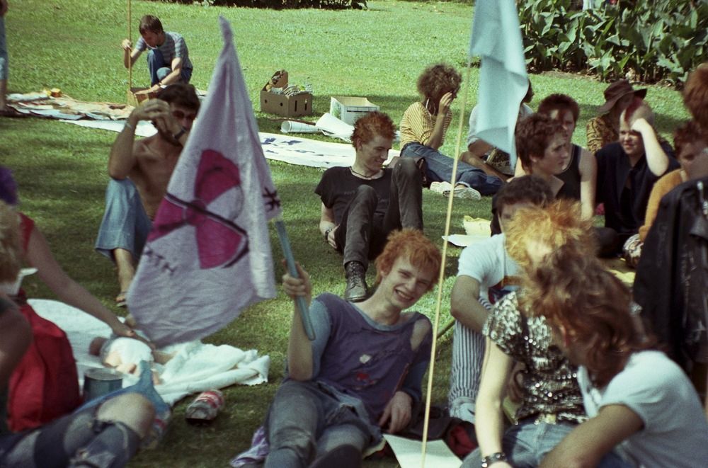 A group of punks lounge on some grass, one is smiling and holding a homemade flag with a peace sign on it.