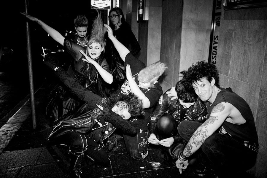 A black and white photo 6 people on a pavement, all in heavy makeup and black clothing.