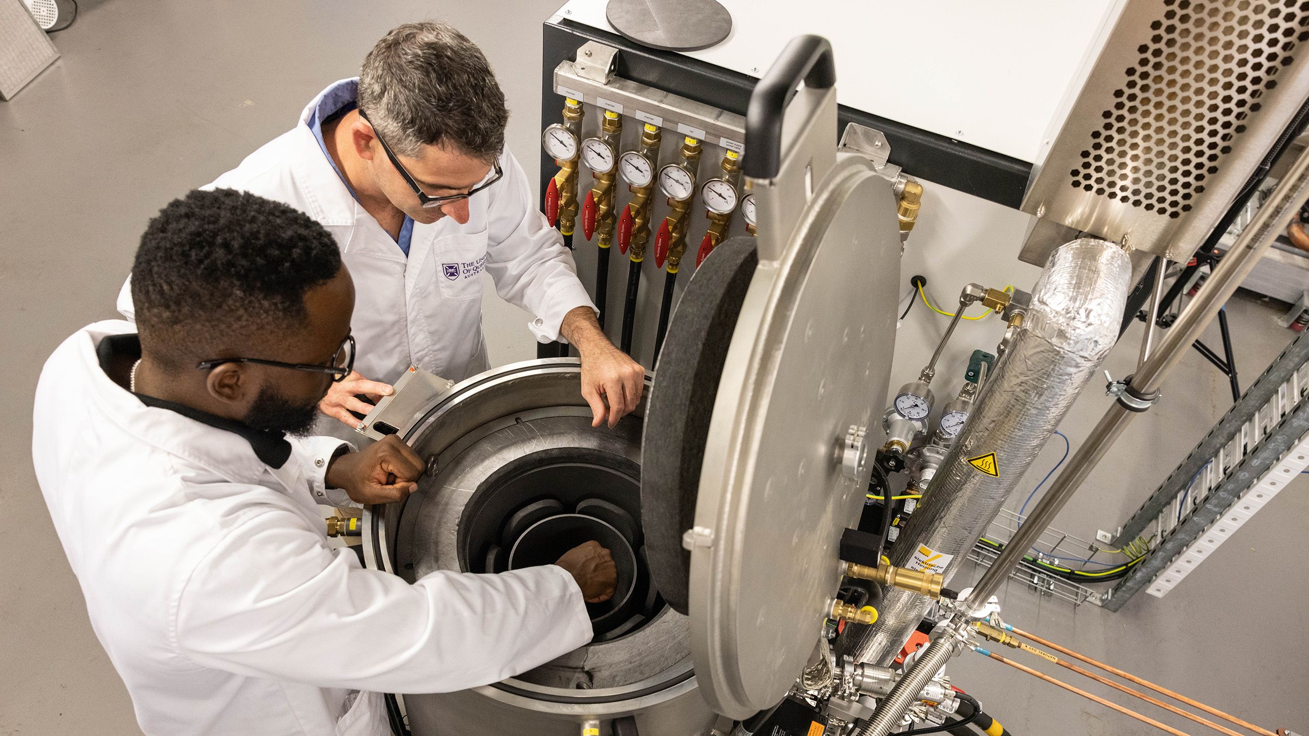 Two people wearing lab coats looking down into a round metal furnace