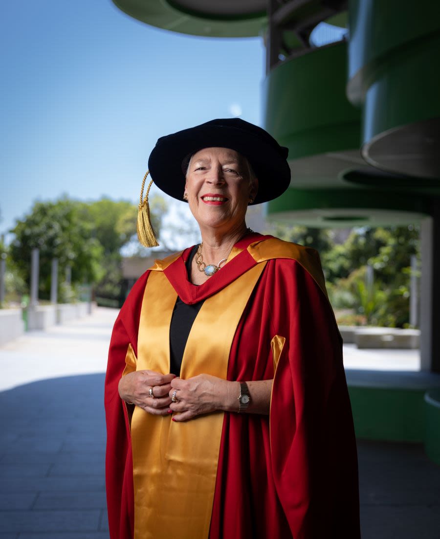 Katherine Hirschfeld standing at the front of the Advanced Engineering Building wearing her red and yellow academic dress and cap.
