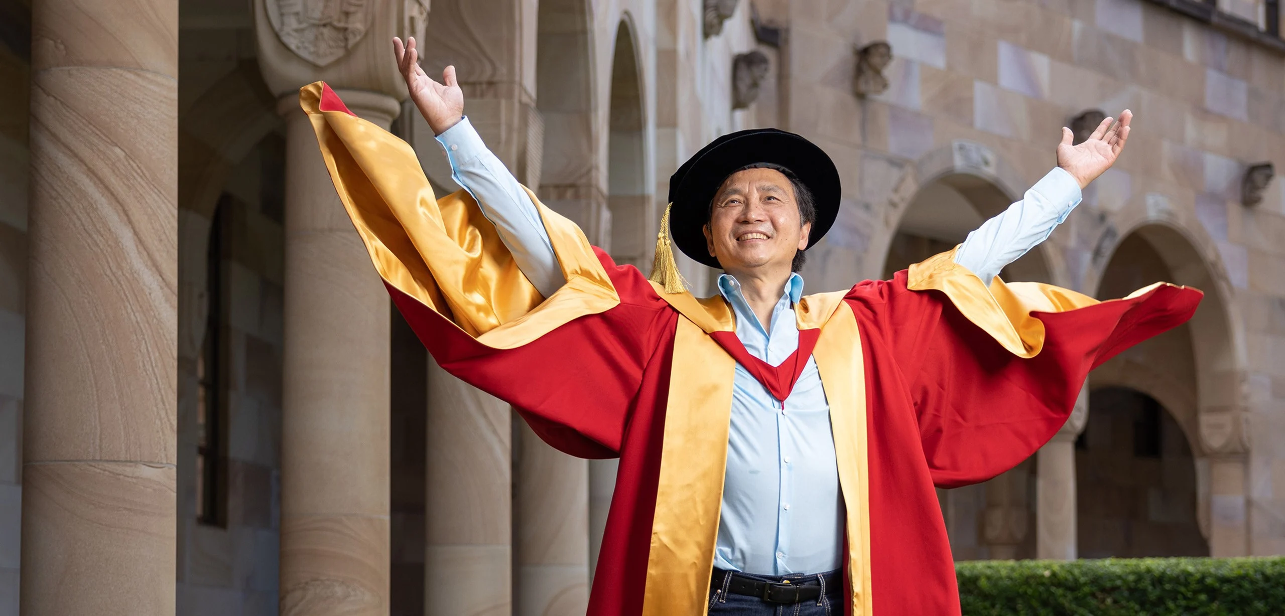 Li Cunxin AO poses in The University of Queensland's Great Court wearing a red and yellow academic dress and cap.
