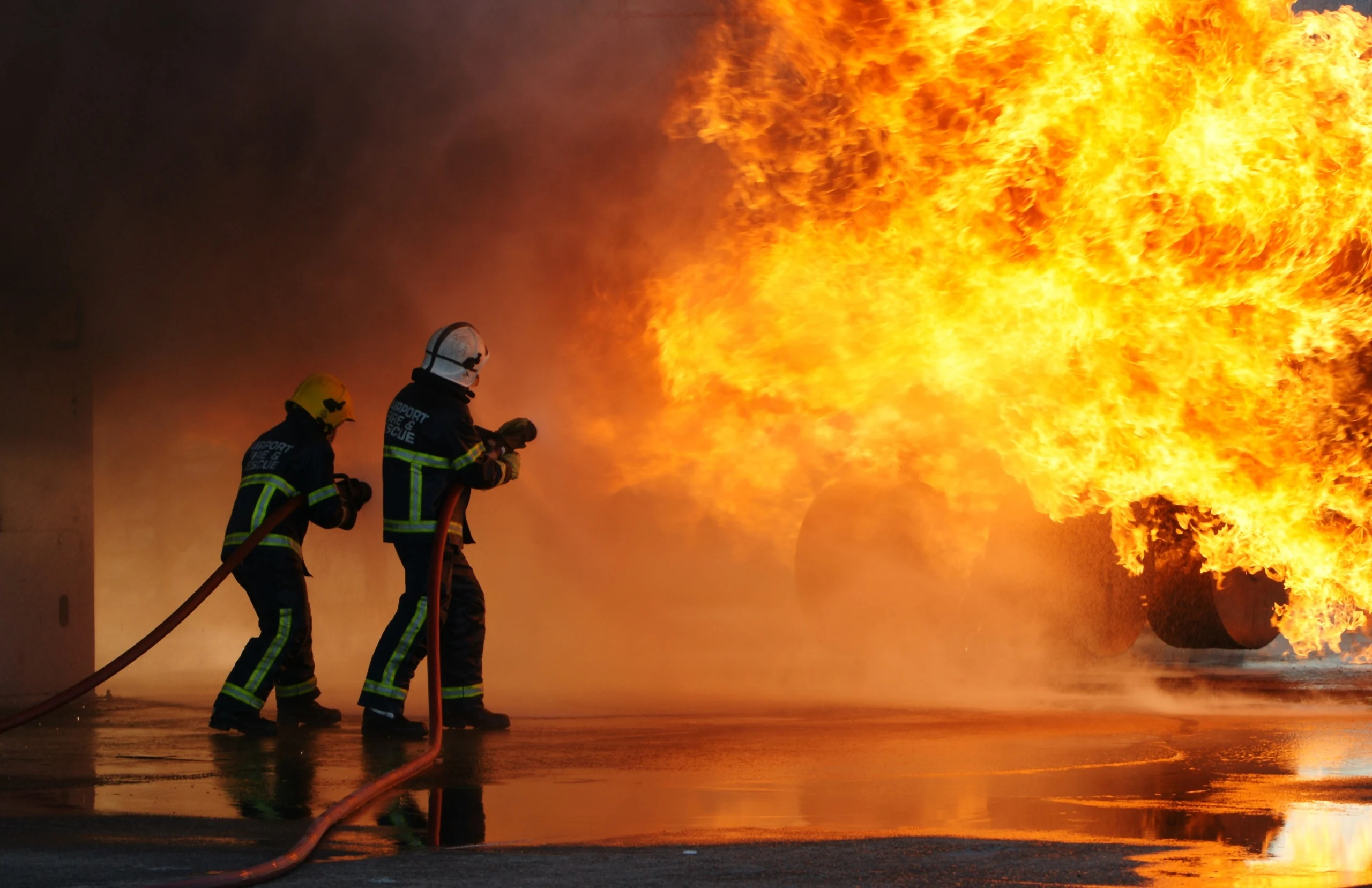 Two people in helmets and dark clothing hold hoses and direct water at flames