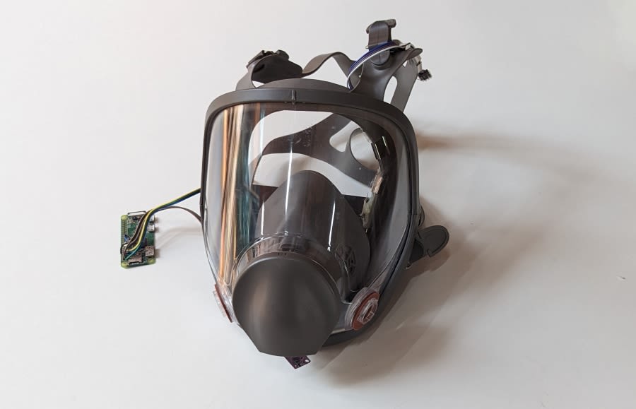 A perspex full face mask with rubber straps and a green circuit board with wires sit on a white background
