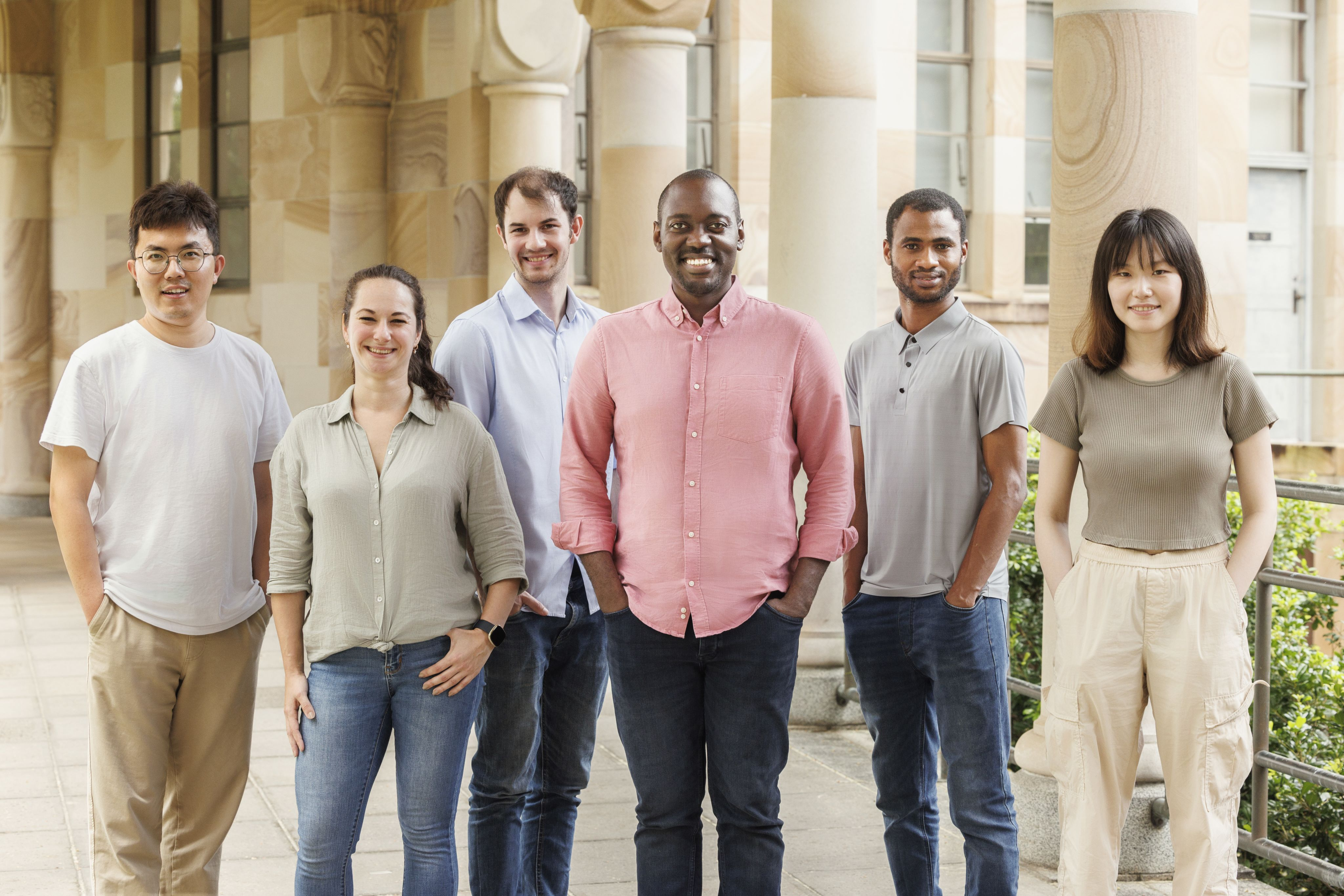 Six people standing smiling at the camera with a sandstone cloister in the background.