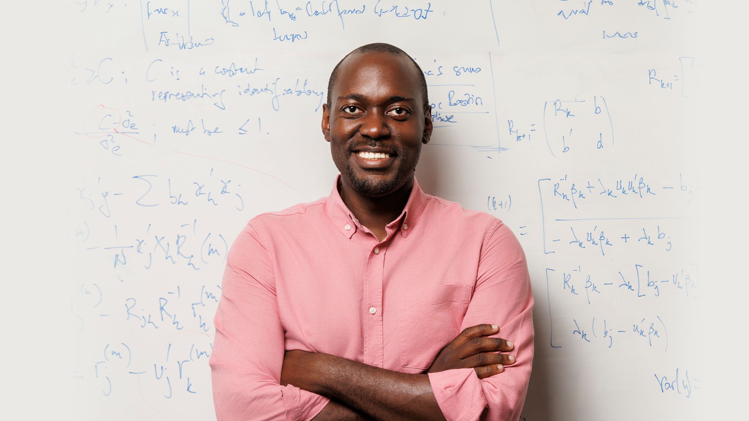 Loic Yengo smiling broadly as he stands with his arms crossed against a whiteboard covered in equations.