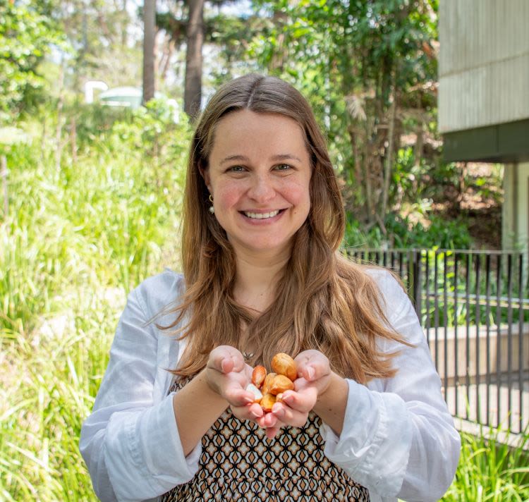 A woman is standing outside, smiling and holding a handful of light brown nuts toward the camera.