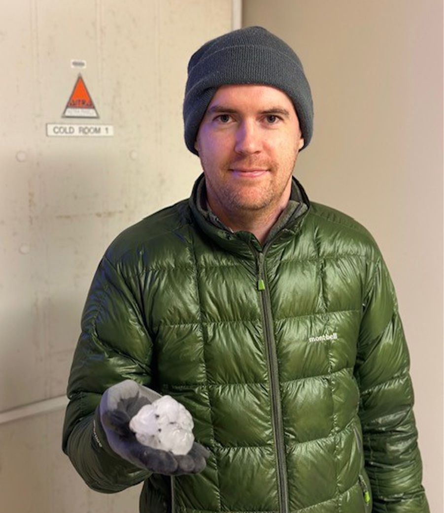 A man in a green puffer jacket and beanie stands holding a hailstone outside a cold room door