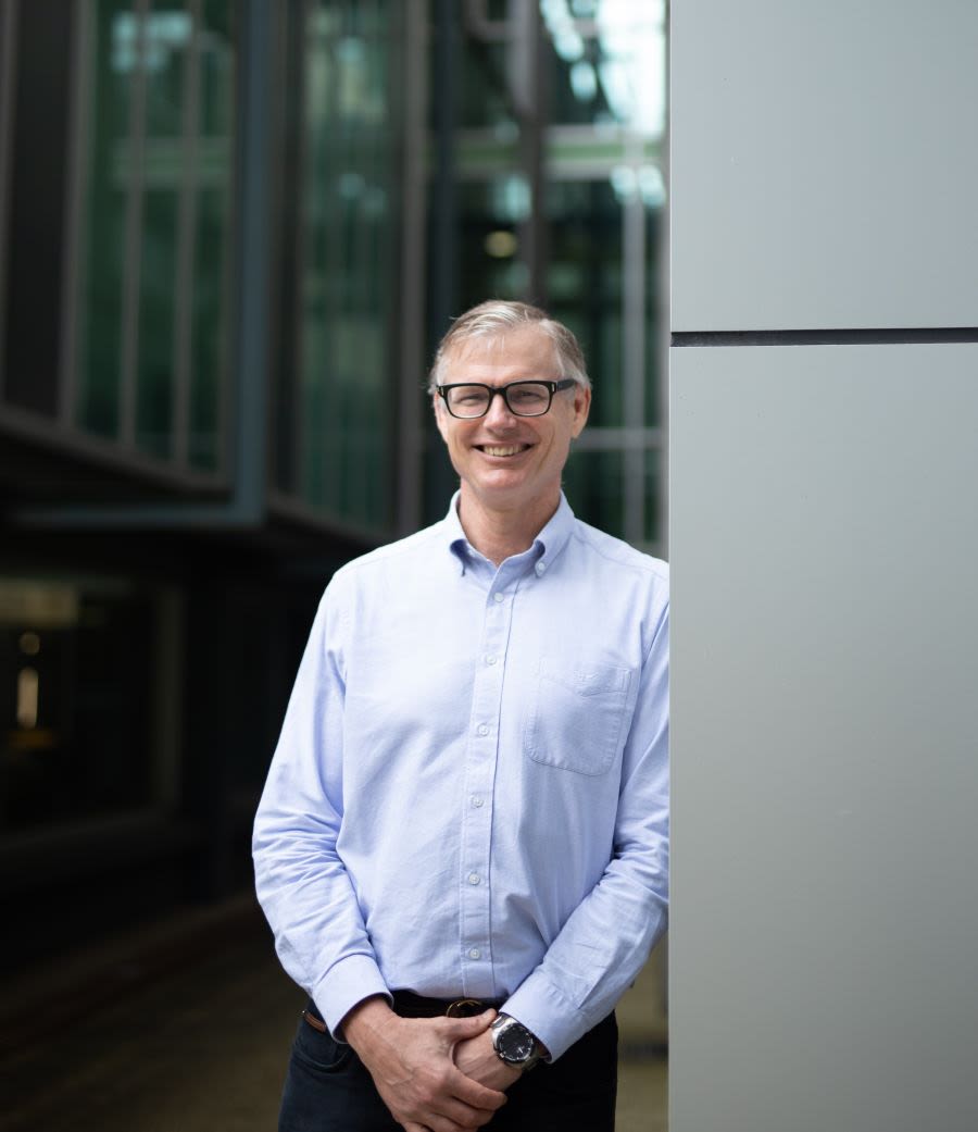 A smiling man in black glasses and a pale blue business shirt leaning against a building outdoors