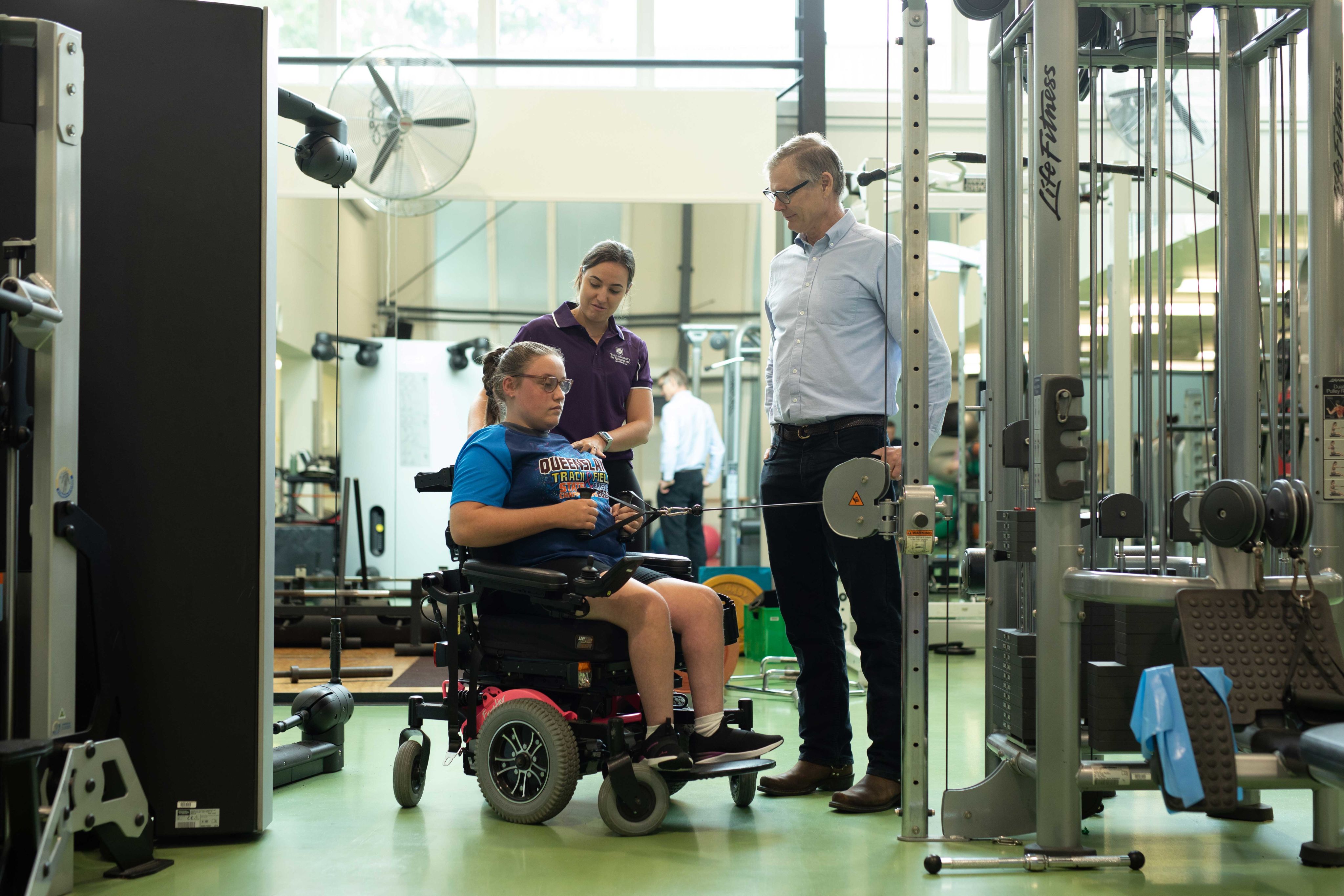 Two trainers guiding a teenage girl, who is using a wheelchair while training in a gym