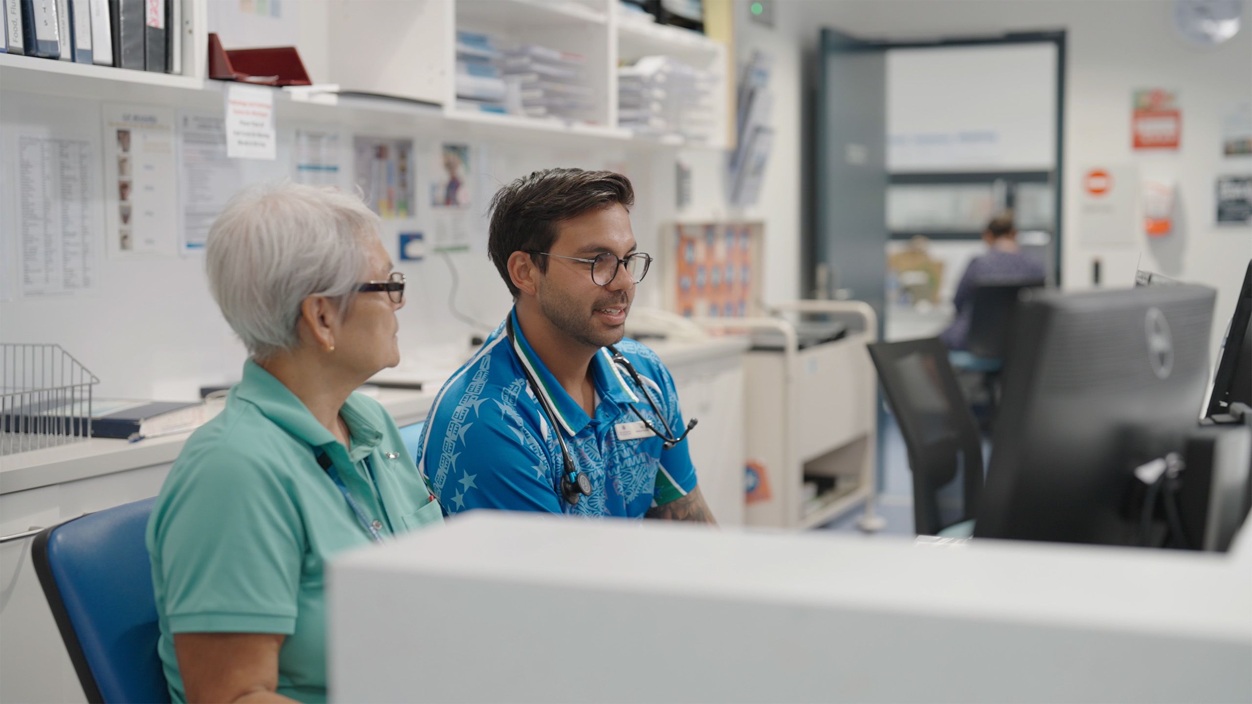 A man and a woman sitting at a hospital reception desk looking at a screen