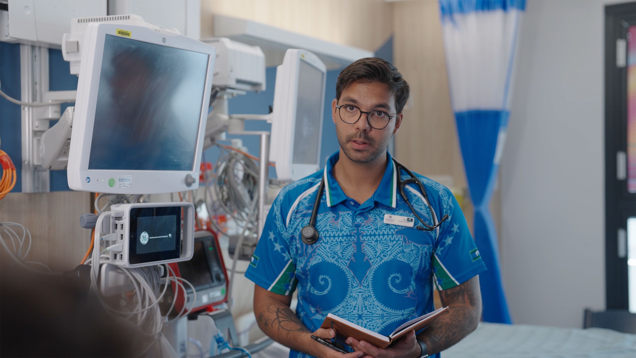 A young man holding a clipboard standing inside a hospital room surrounded by equipment
