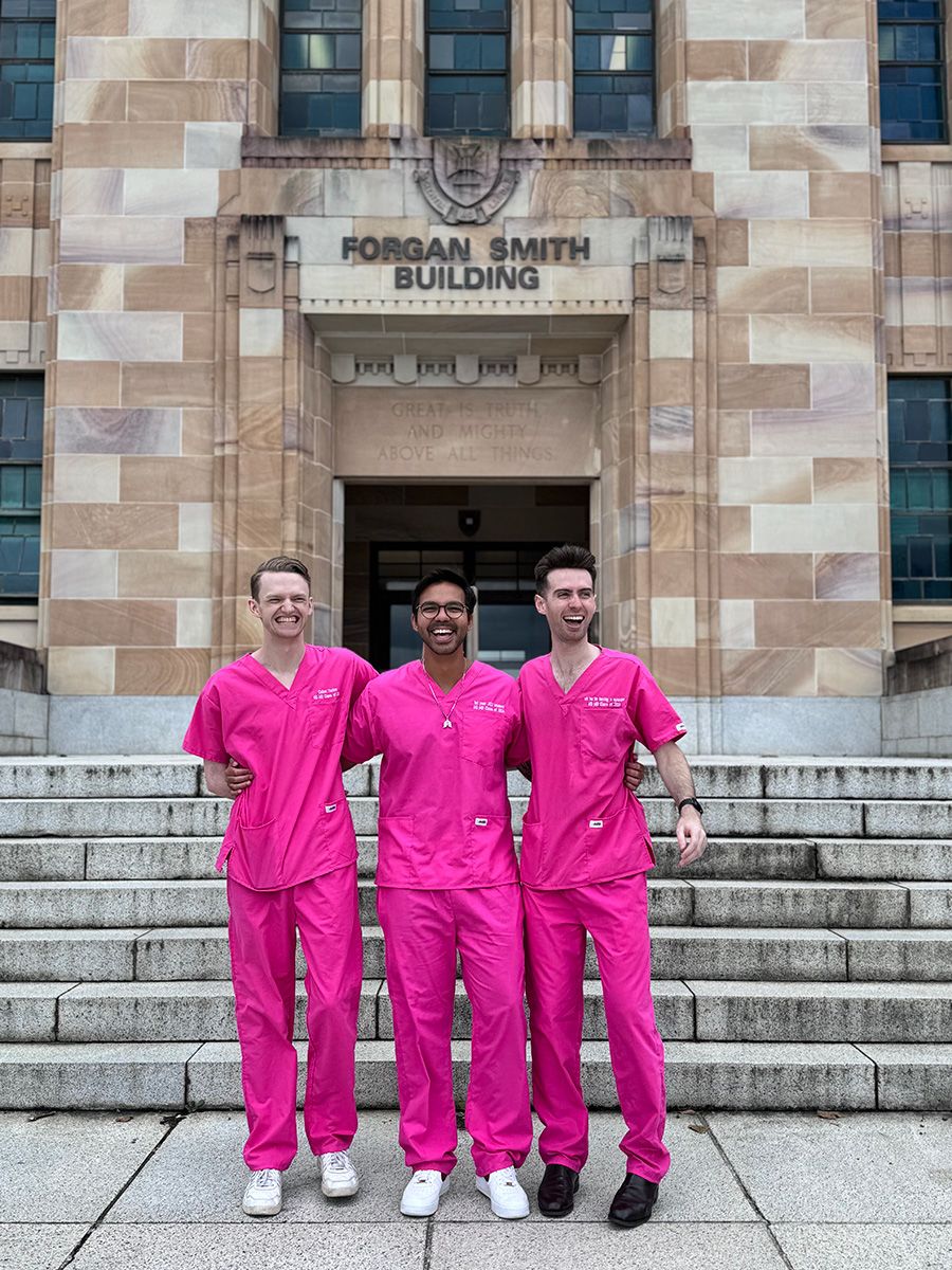 Three young men in pink scrubs standing outside a standstone building