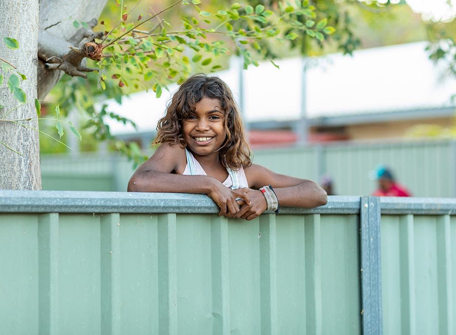An Indigenous child resting their arms on a tall metal fence and smiling