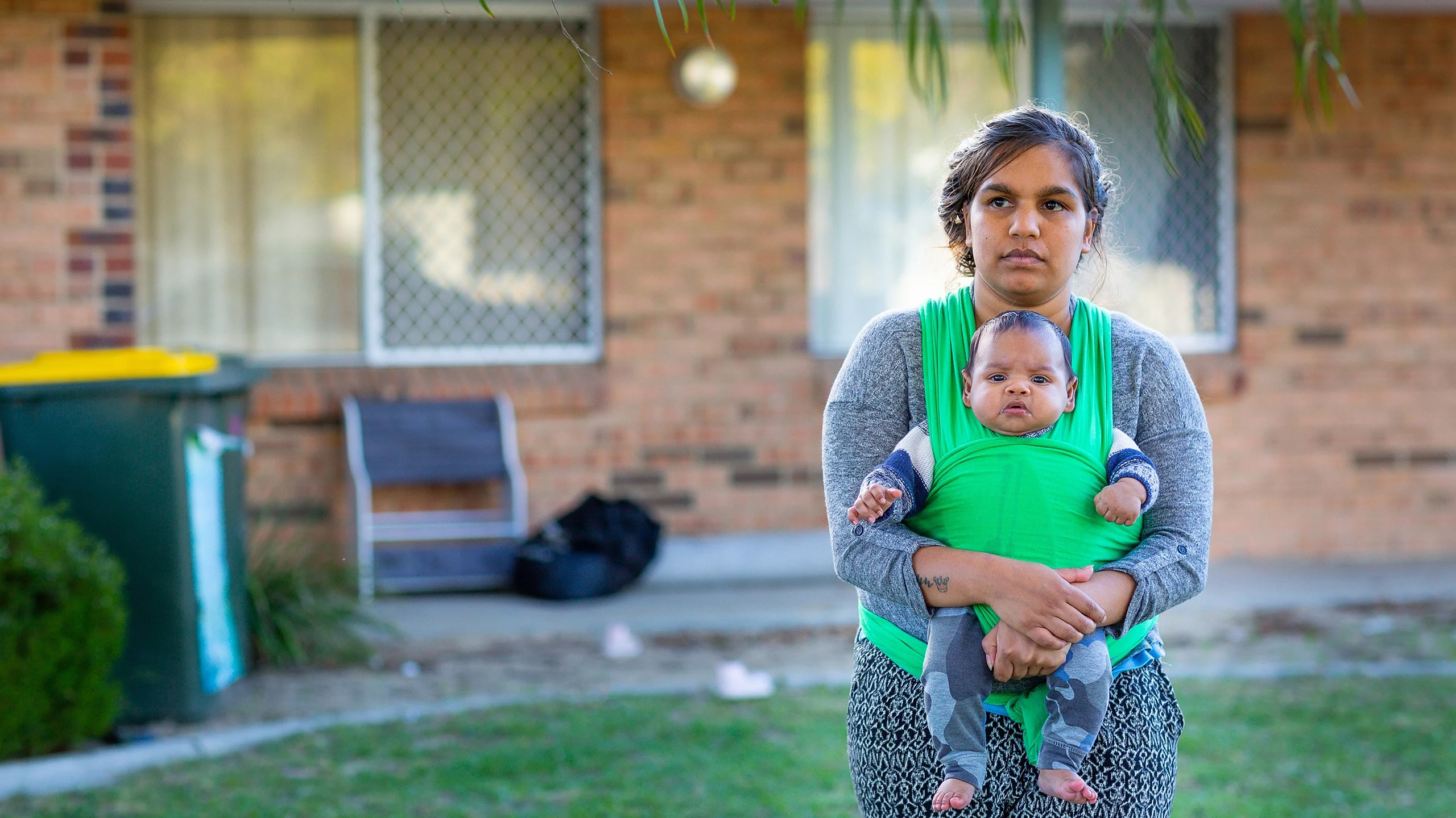 An Indigenous woman standing in front of a brick house. A baby is secured to her chest with a bright-green sling.