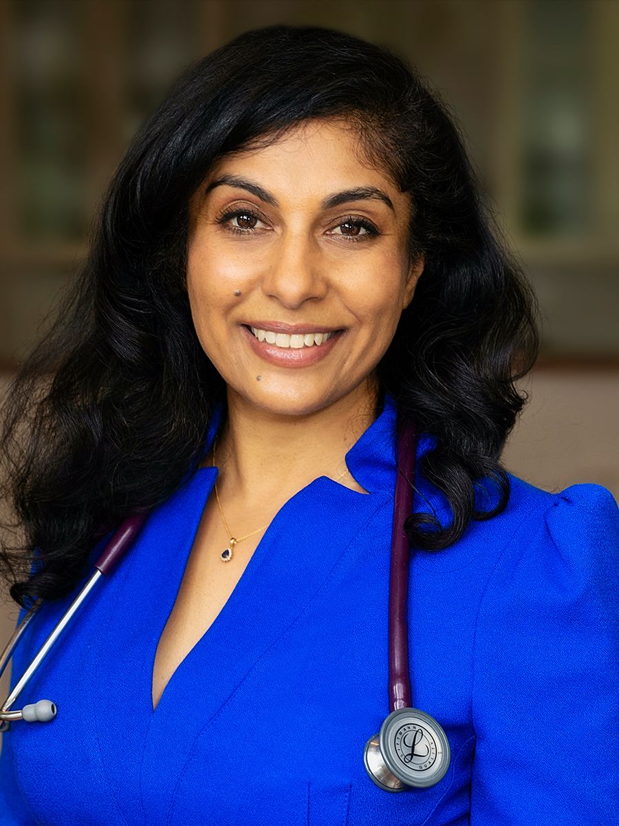 A headshot of Dr Niroshini Kennedy smiling while wearing blue scrubs. A stethoscope is draped around her neck.