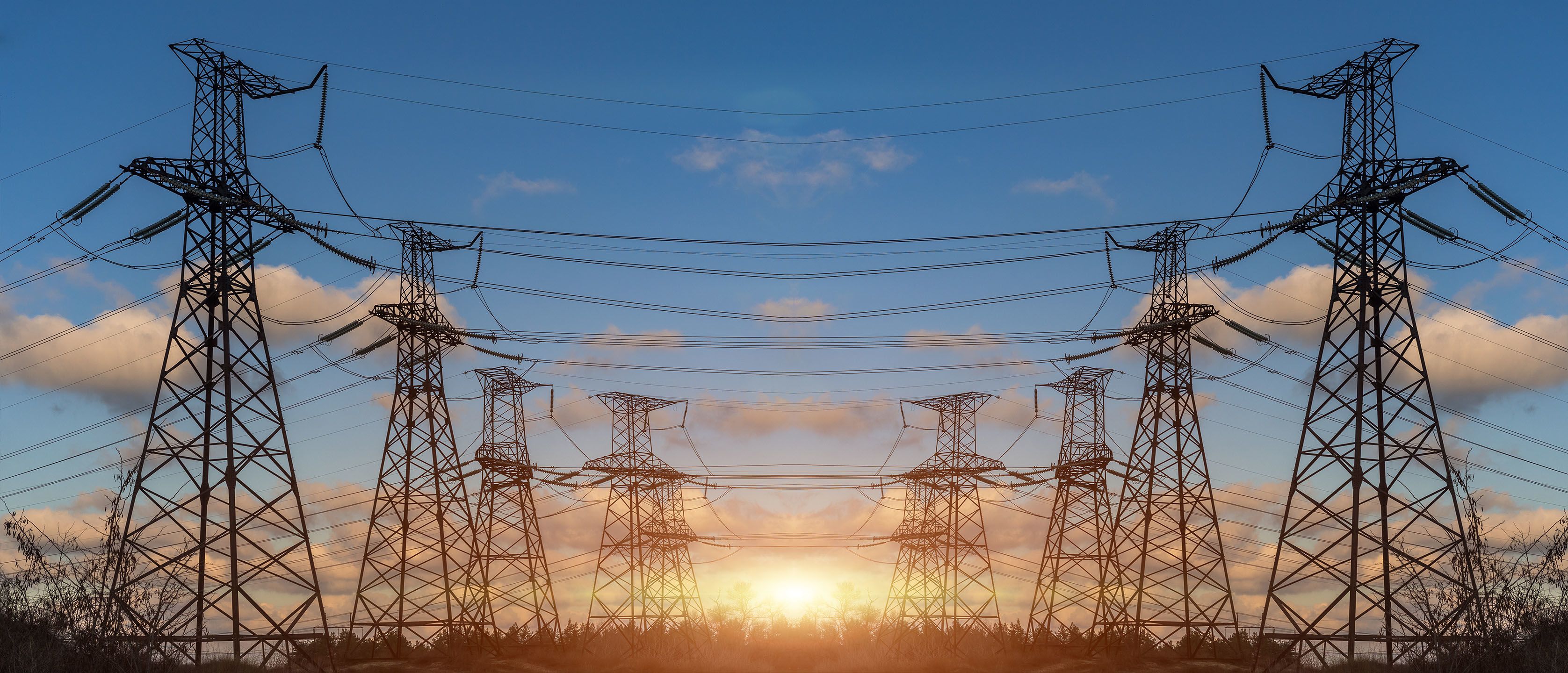High voltage electric transmission towers silhouetted against a blue sky and rising sun. 