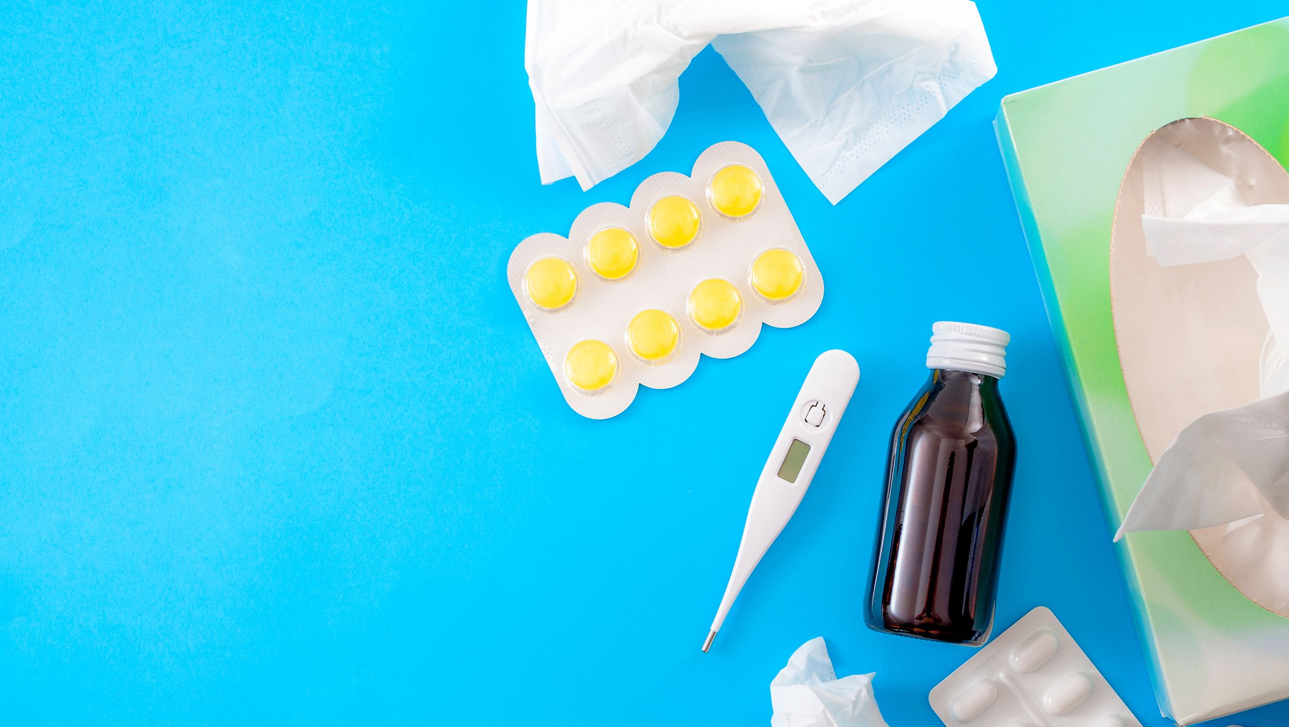 A sheet of yellow lozenges, an electronic thermometer, a bottle of unlabelled medicine, and tissues laying on a blue surface