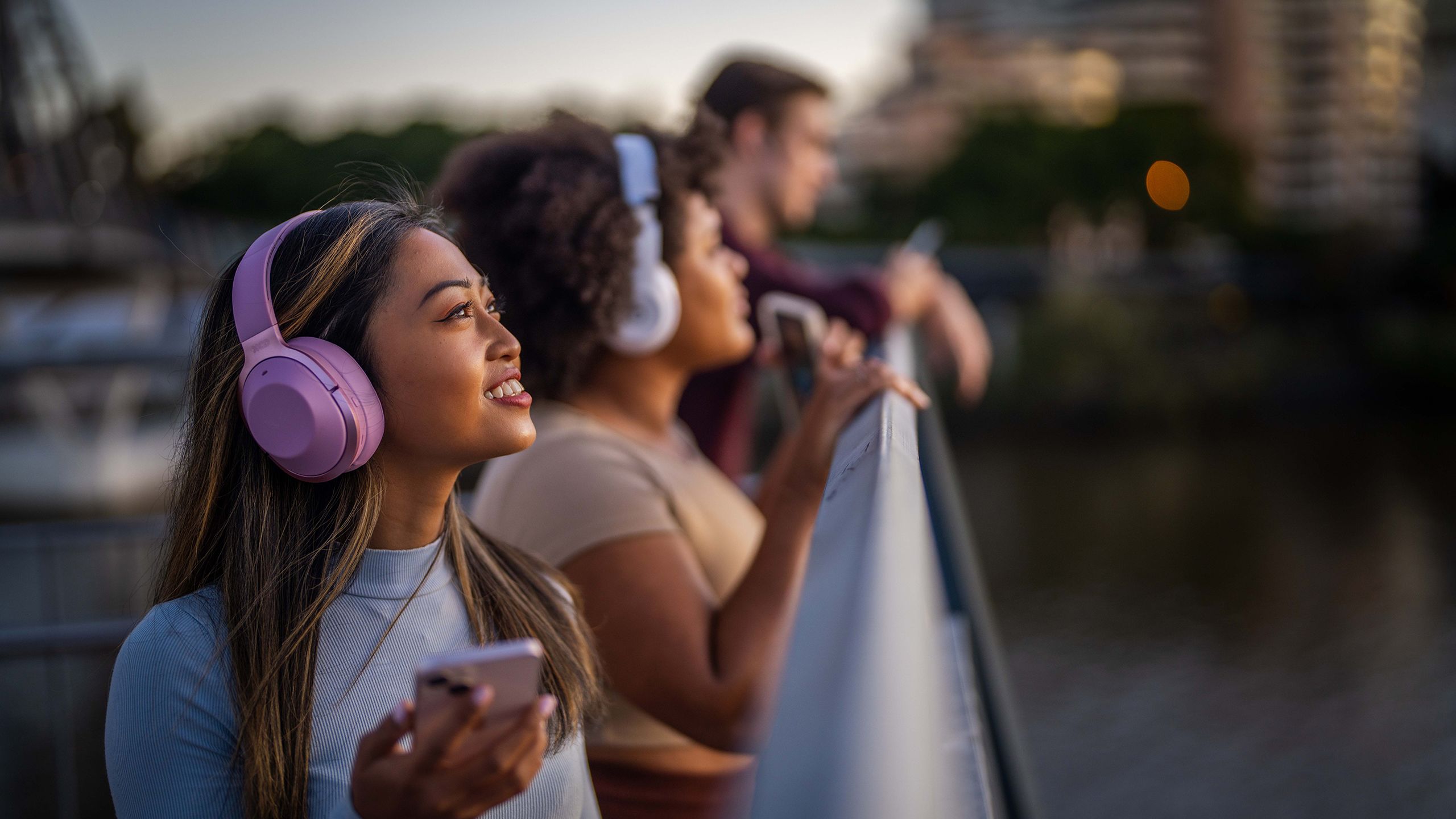 Three people standing on a bridge overlooking a river. They are wearing headphones and are each holding a mobile phone in their hands.