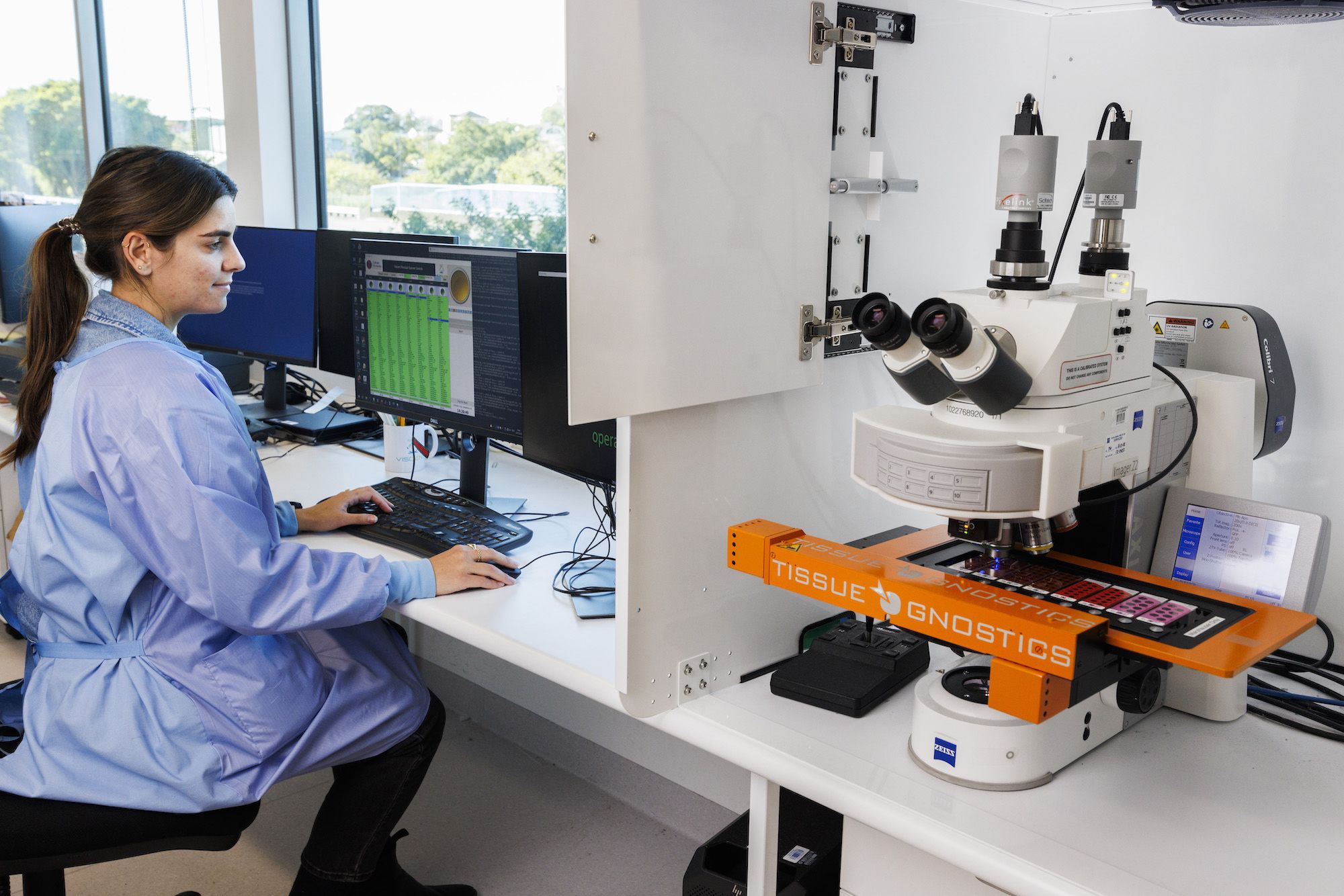 A female scientist in a blue coat sitting in front of a computer screen and next to a digital scanner