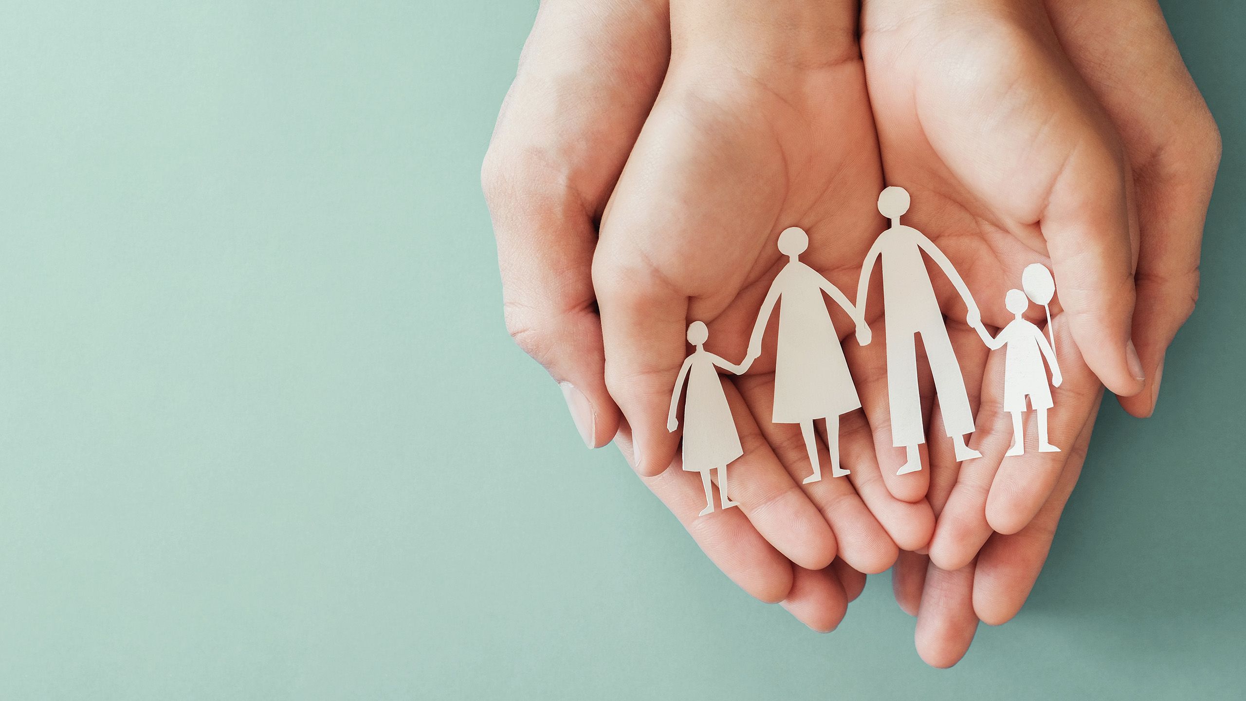 A pair of hands cupping a smaller pair of hands. A white cardboard cutout of a family with a mother, father, and two children is resting in the smaller hands.