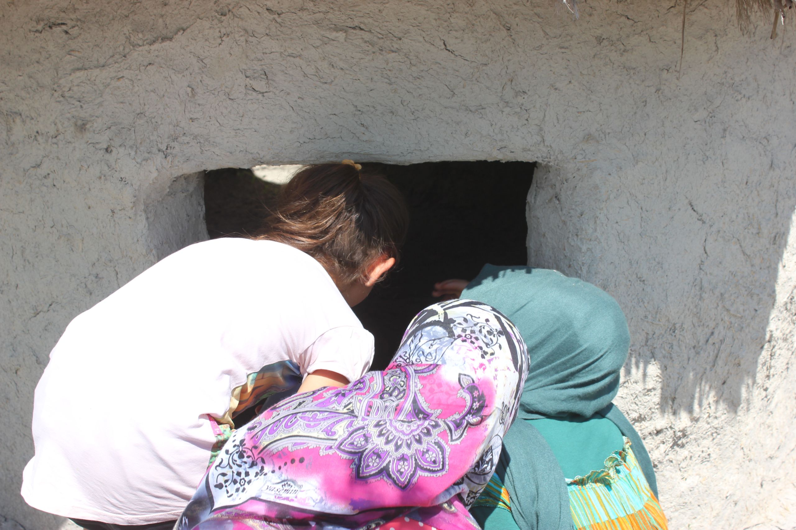 Three children peer inside the window of a mud brick hut