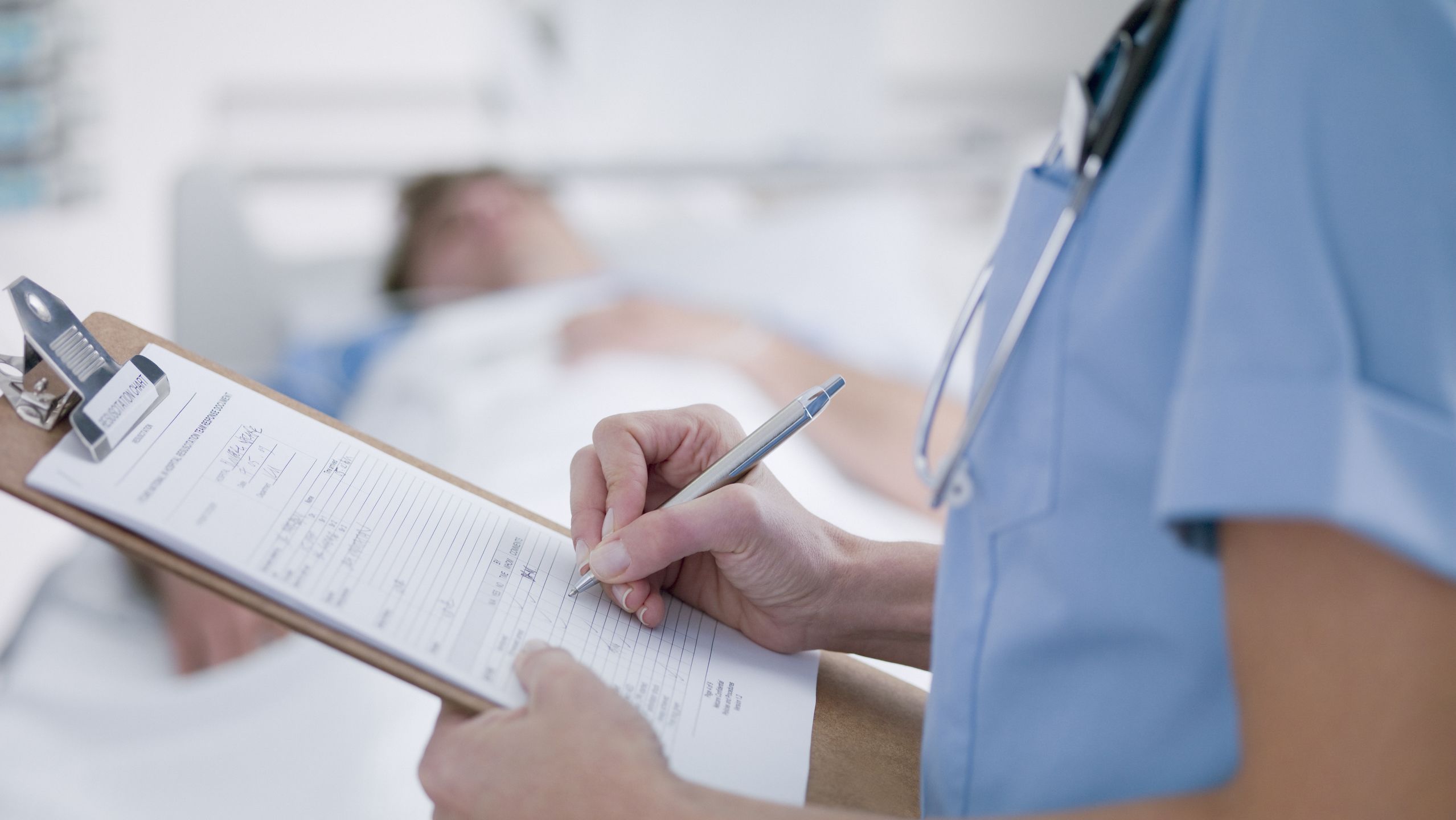 A female nurse in blue scrubs looks down at a hospital chart examining information. Behind them, a male patient is resting in a hospital bed.