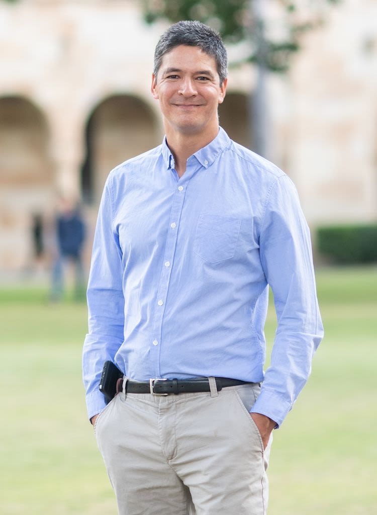 Professor Mark Horswill, wearing beige pants and a light blue buttoned shirt, with his hands in pockets, standing on UQ's Great Court.