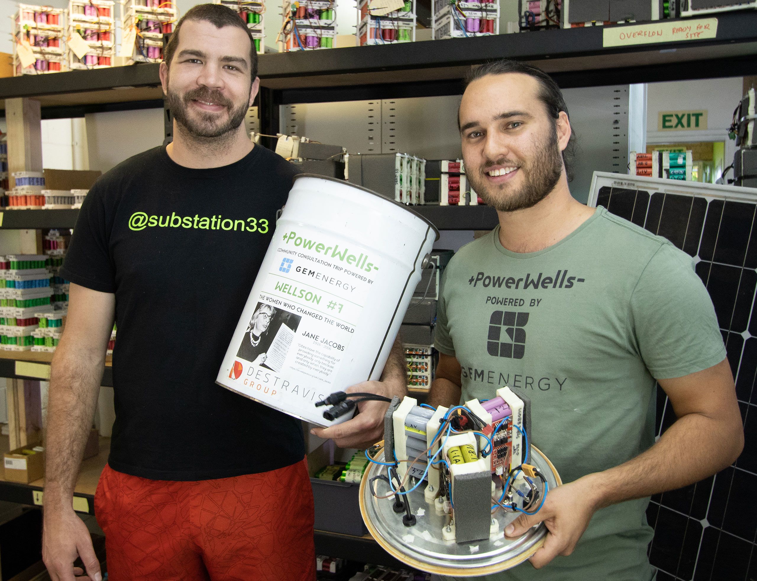 Nicholas and Bradley showing off their PowerWell. A shelf of electronics is behind them. 