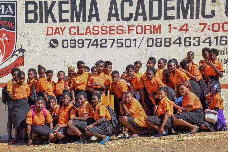 A group shot of Bikema Academy School girls - they're dressed in orange tops and grey skirts.