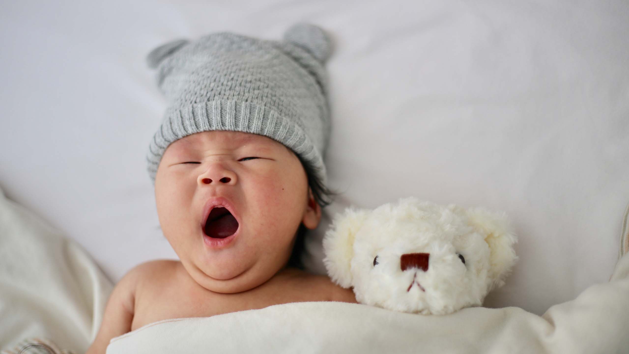 A yawning baby wearing a grey knit hat with ears. It is tucked in to a bed and has a white teddy bear tucked in next to it.