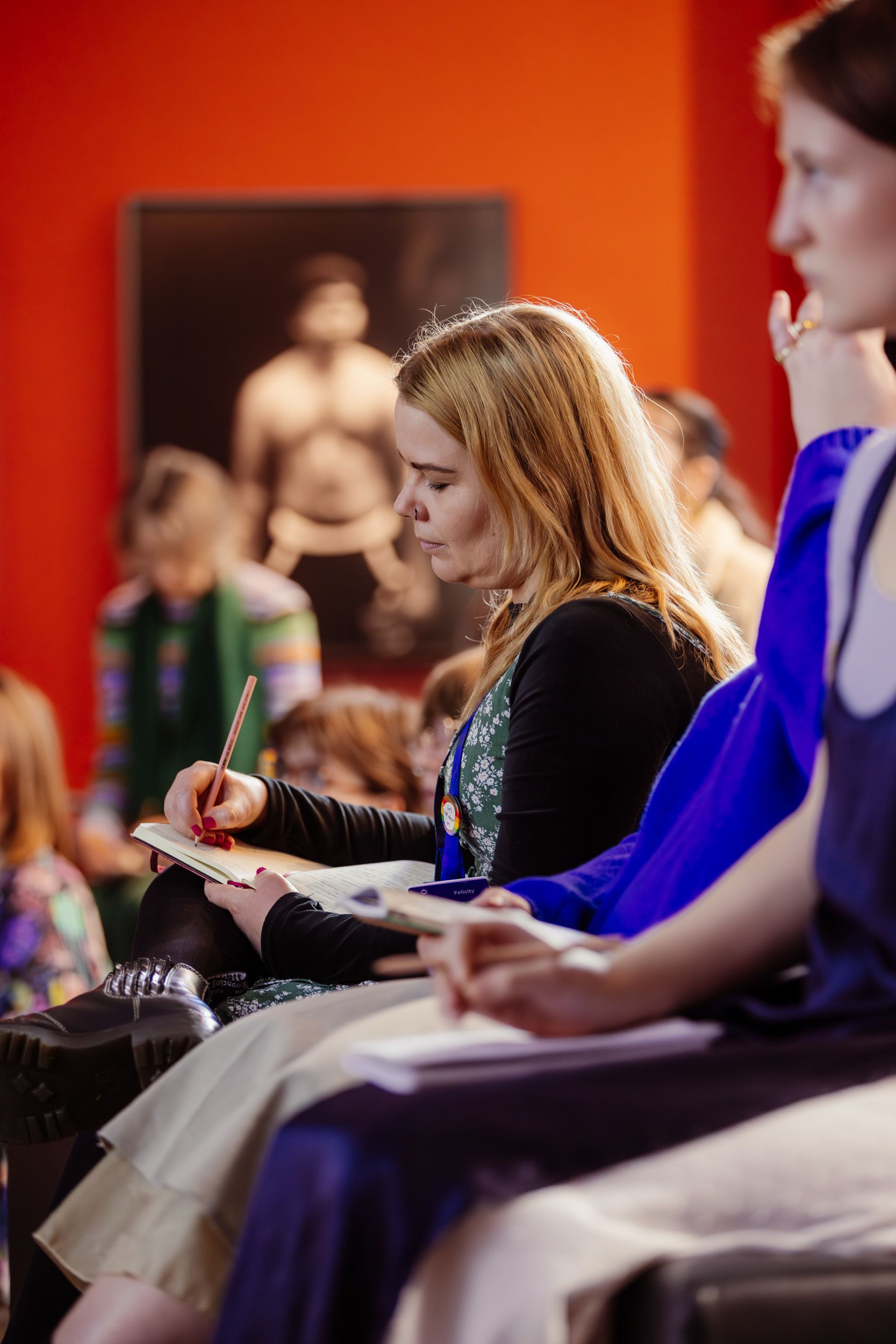 A young person is writing in a notebook with a pencil. There are people sitting on either side of them. 